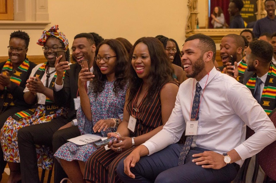 Group of individuals sitting on chairs and facing the left, appearing to laugh, some of them with their phones out.