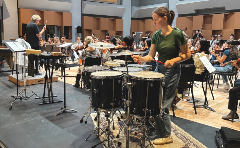 A student orchestra rehearsal or performance in a concert hall, with a conductor leading musicians across strings, winds, brass, and percussion sections. 
