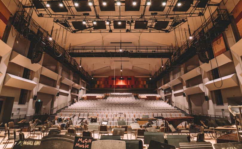 A music performance hall viewed from stage, showing tiered seating, balconies, orchestra chairs, music stands, instruments, and a conductor area.