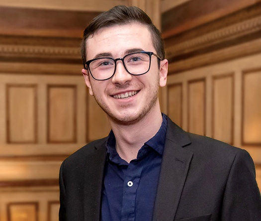 A man wearing a dark blazer and navy-blue shirt standing indoors against a wooden paneled wall.