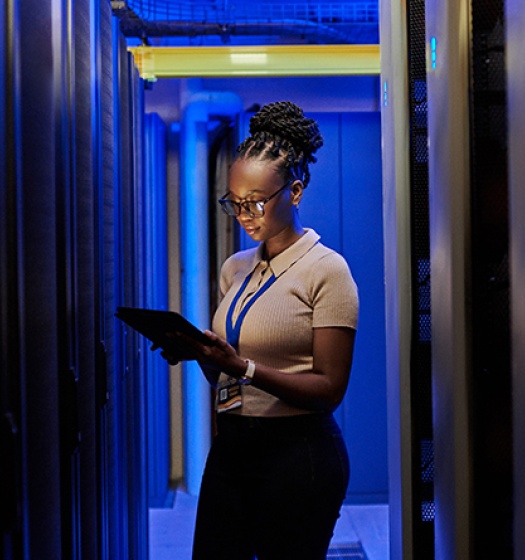  A woman standing between server racks in a data center, holding a tablet and wearing a lanyard badge. 