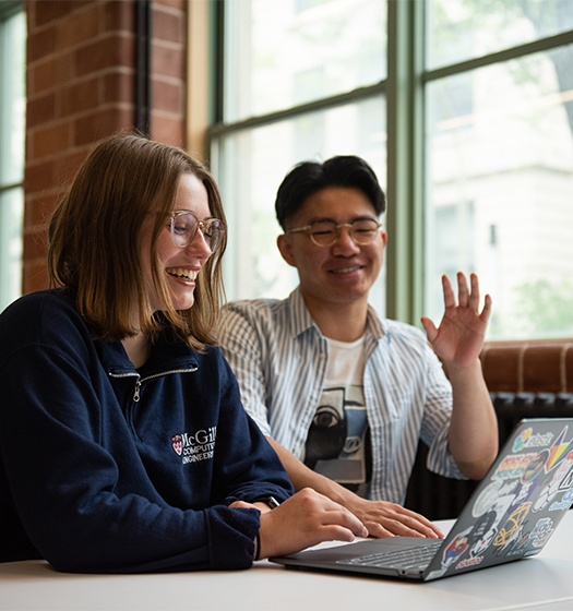 Two students sitting at a table with a laptop covered in colorful stickers, engaged in discussion or collaboration. 